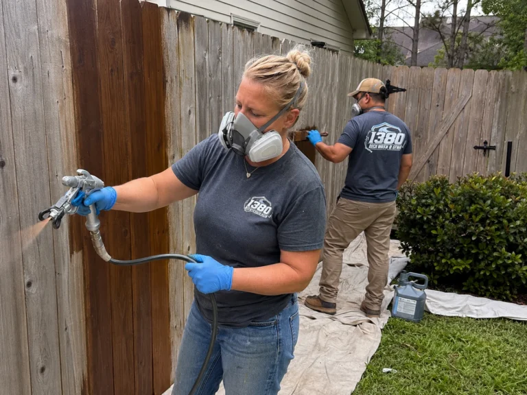 Worker spraying stain onto a wood privacy fence while wearing protective equipment
